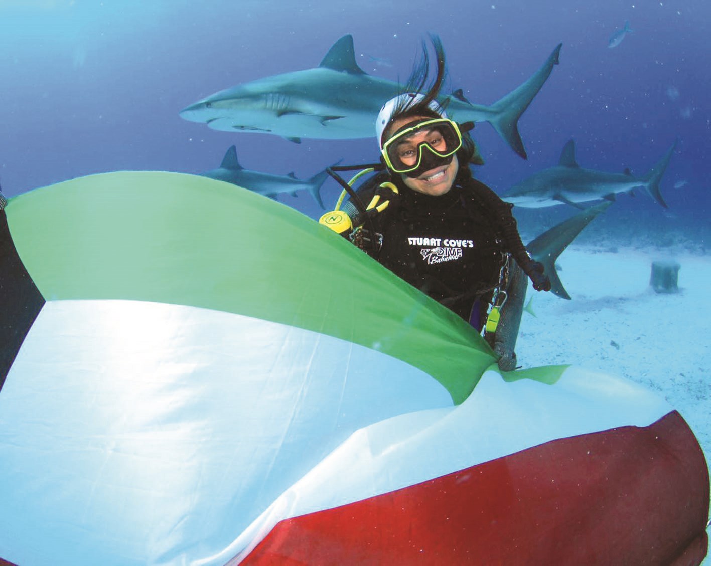 ابتسامة مع علم الكويت في الأعماق وخلفها أسماك القرش